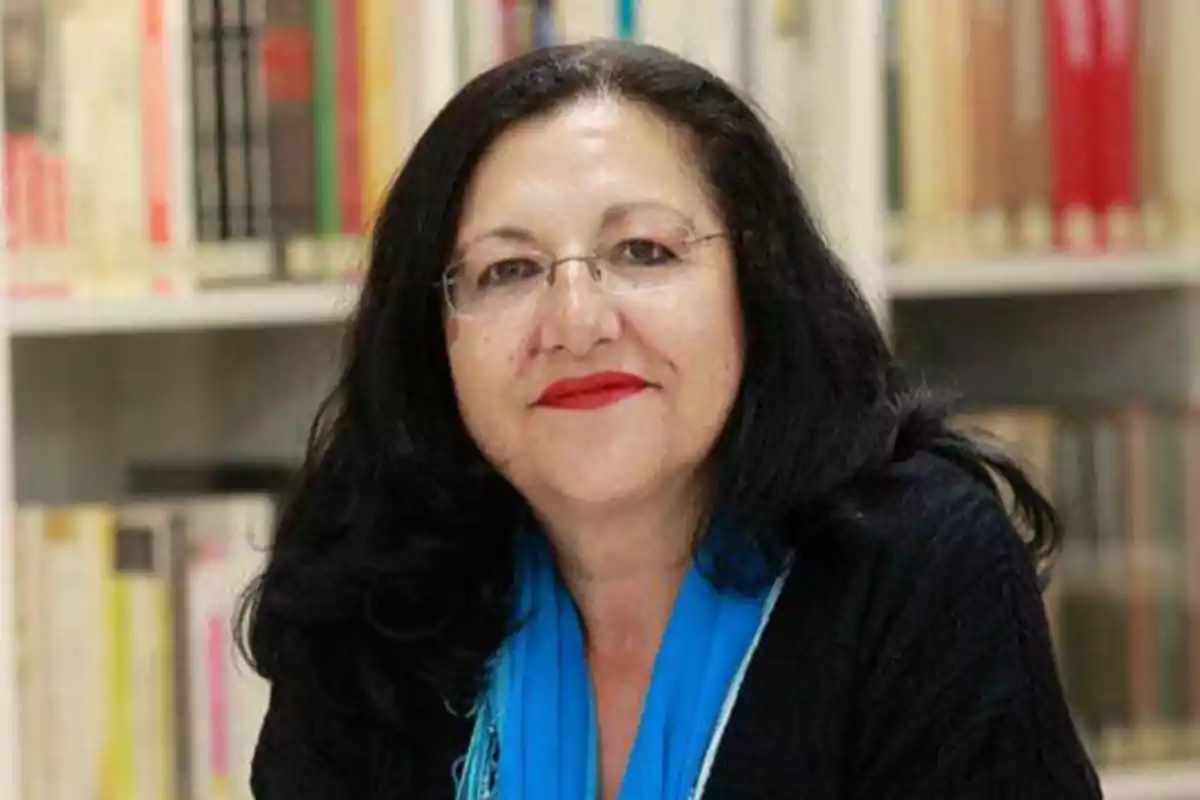 Mujer con cabello oscuro y gafas, sonriendo, con una bufanda azul, frente a una estantería de libros.