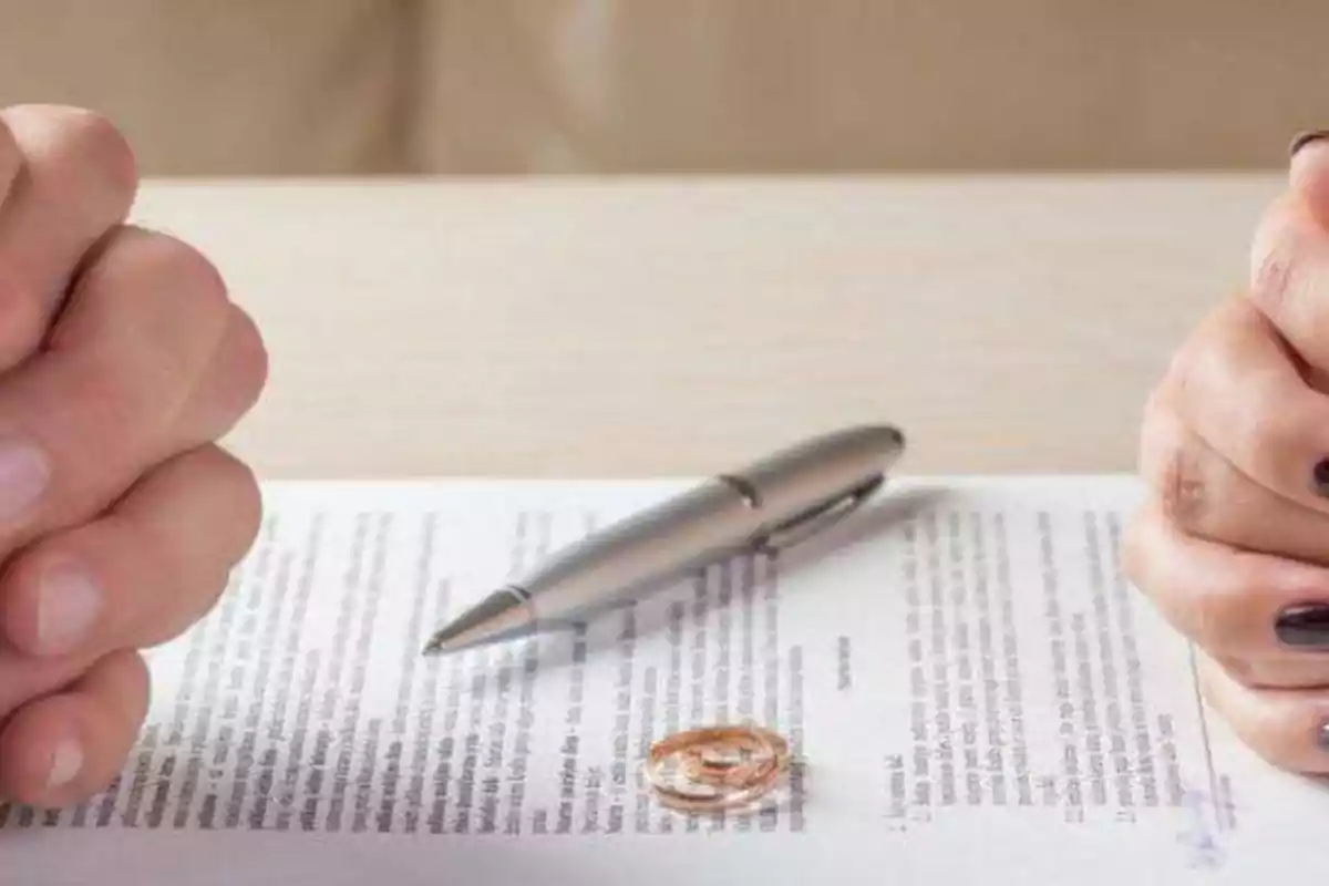 Dos manos enfrentadas con un documento, un bolígrafo y dos anillos sobre la mesa.