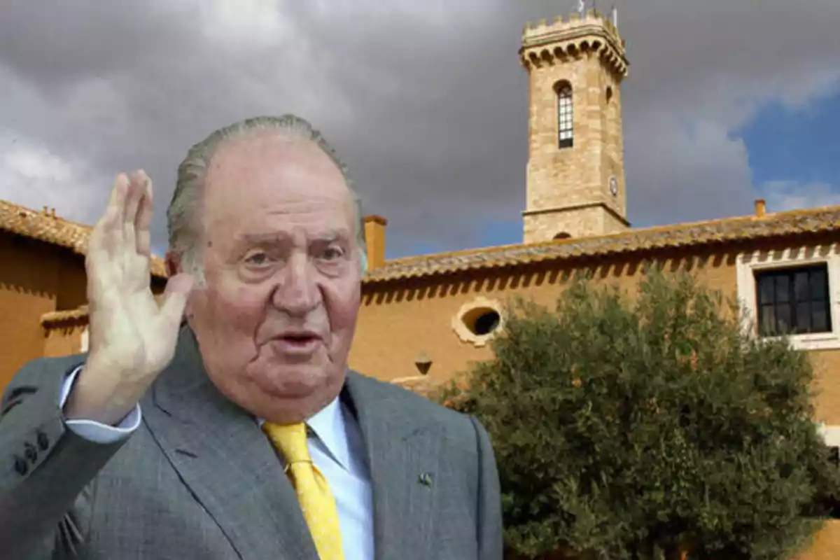 Un hombre mayor con traje y corbata amarilla levantando la mano frente a un edificio de estilo rústico con una torre y cielo nublado.
