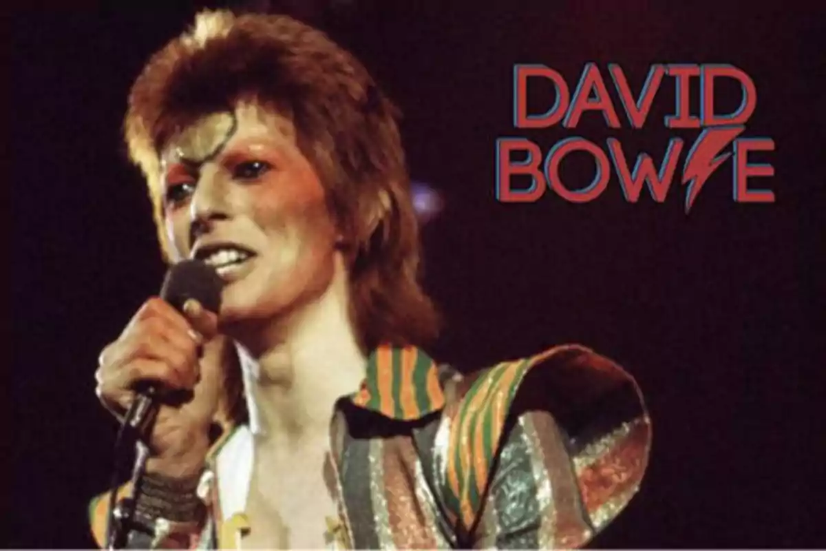 Persona con maquillaje llamativo y vestimenta colorida sosteniendo un micrófono con el texto "David Bowie" en el fondo.