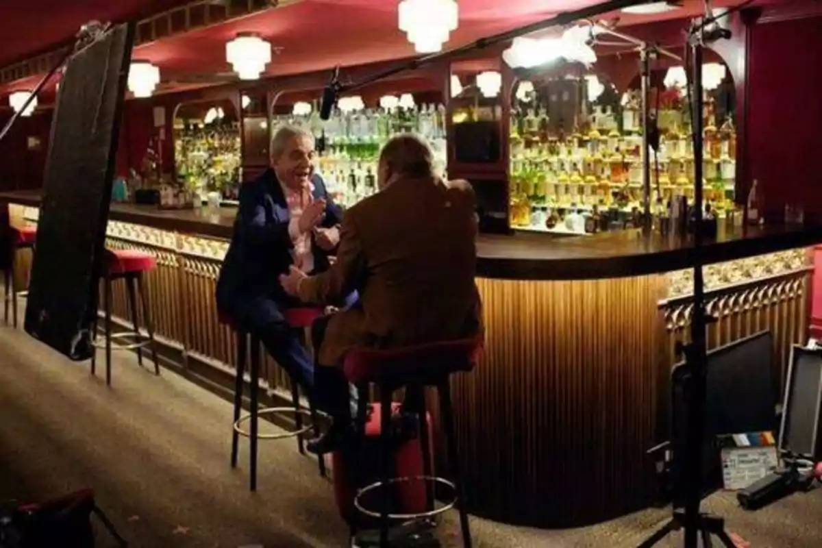 Dos personas conversan animadamente en un bar bien iluminado, con equipo de grabación visible alrededor.