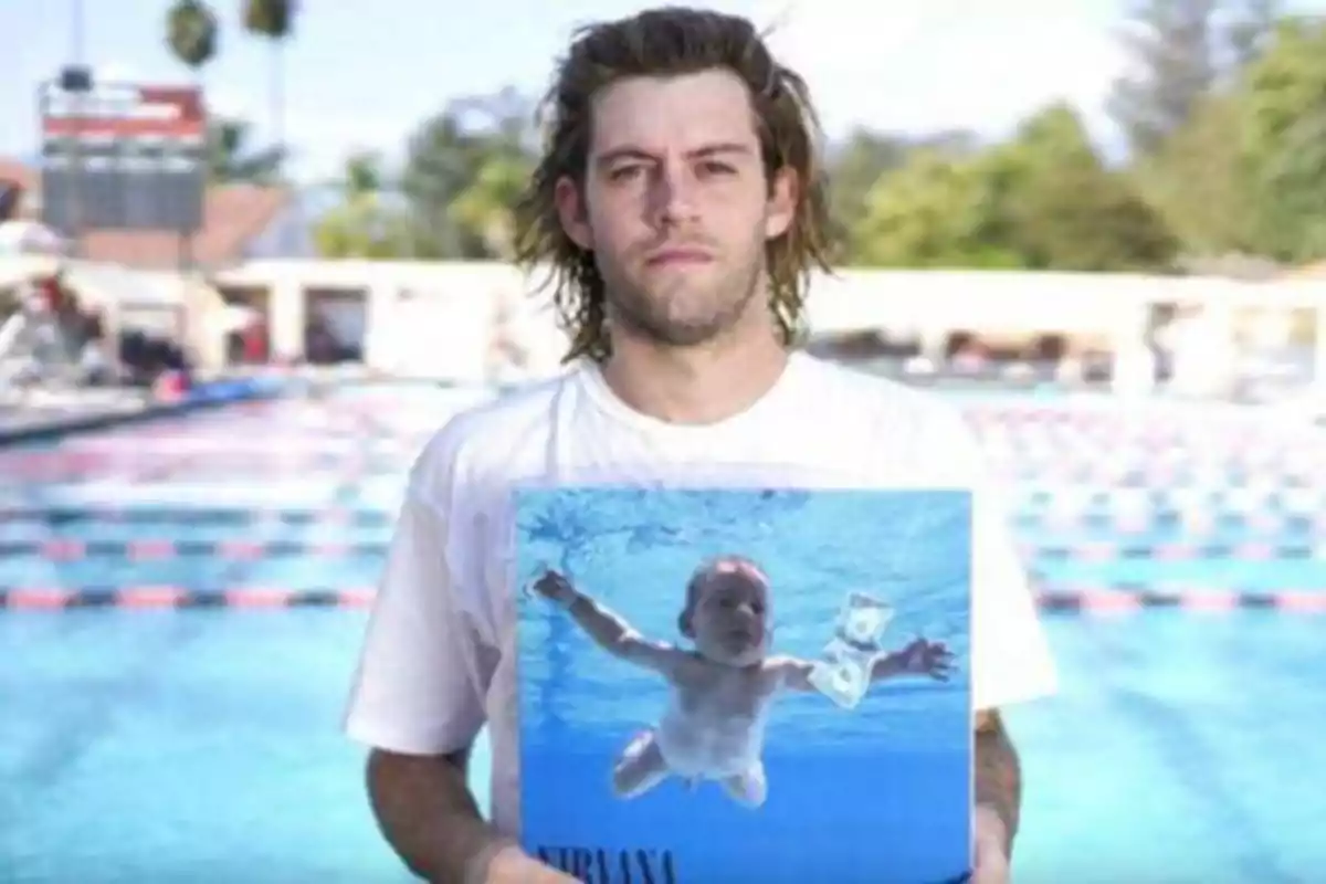 Un hombre con cabello largo y barba ligera sostiene un álbum de Nirvana frente a una piscina.