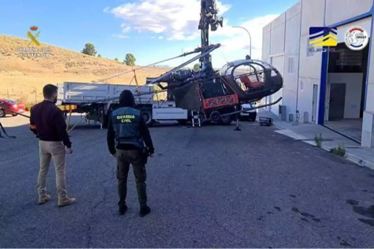 Dos personas observan un helicóptero militar en un remolque, con logos de la Guardia Civil y Europol en la imagen.