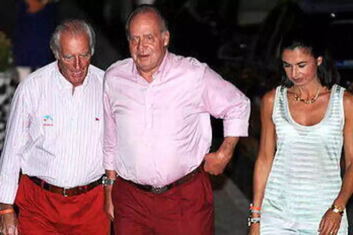 Tres personas caminando juntas, dos hombres mayores con camisas claras y pantalones rojos, y una mujer con un vestido sin mangas a rayas.