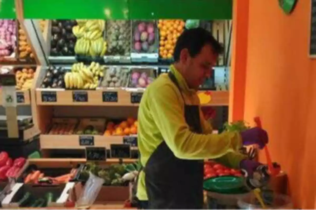 Un hombre con guantes morados y un delantal negro está trabajando en una frutería, rodeado de frutas y verduras.