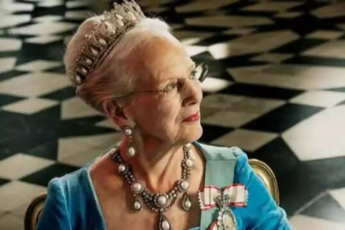 Una mujer mayor con una tiara y joyas elegantes, vestida con un traje azul, sentada en una habitación con un suelo de baldosas en blanco y negro.