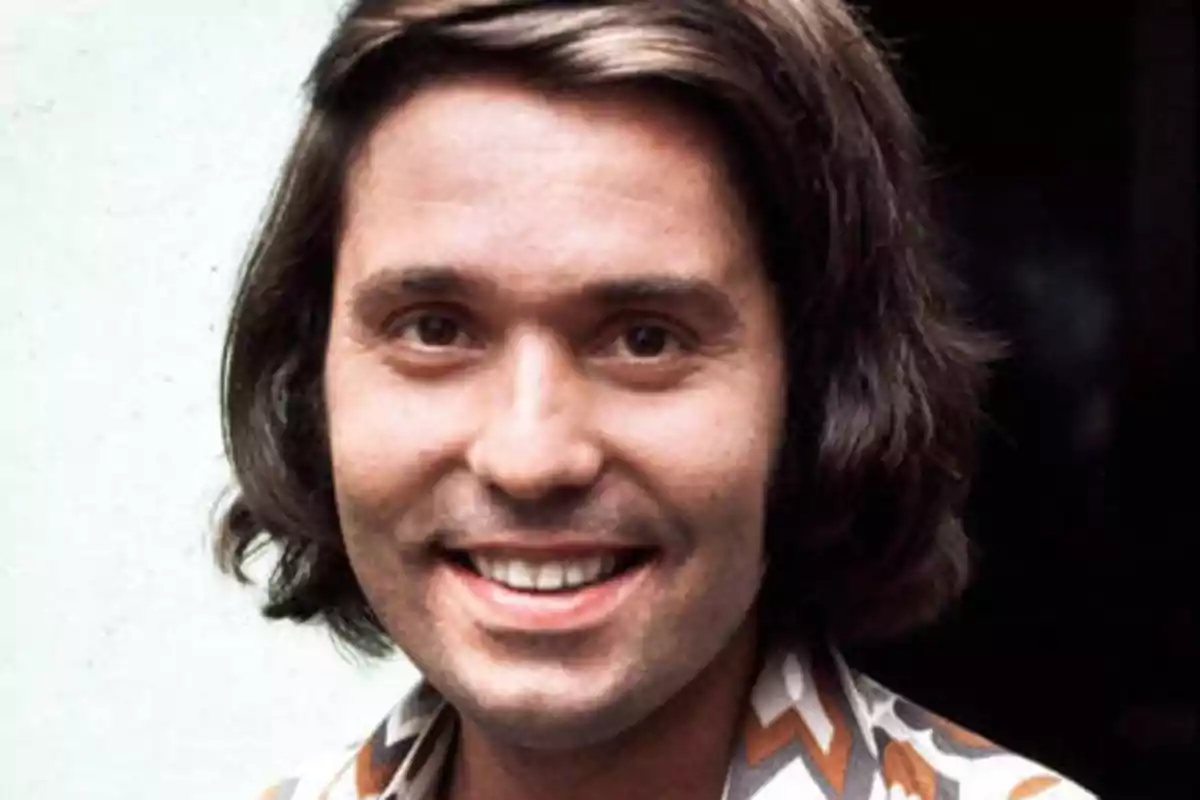 Un hombre con cabello largo y oscuro, sonriendo, con una camisa de patrones geométricos.