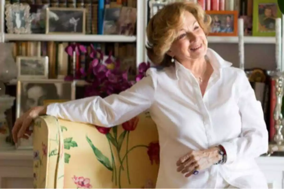 Una mujer mayor sonriente, vestida con una camisa blanca, está sentada en un sillón amarillo con estampado floral, con una estantería llena de libros y fotos en el fondo.