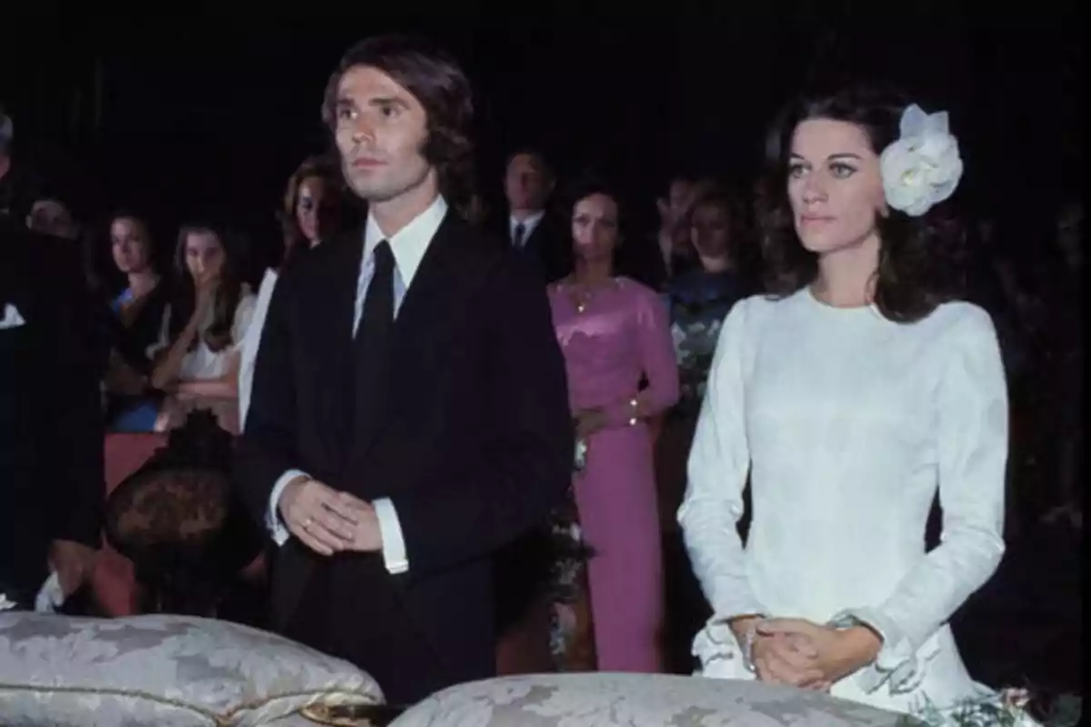 Una pareja vestida formalmente, el hombre con traje oscuro y la mujer con un vestido blanco y una flor en el cabello, en lo que parece ser una ceremonia con varias personas al fondo.