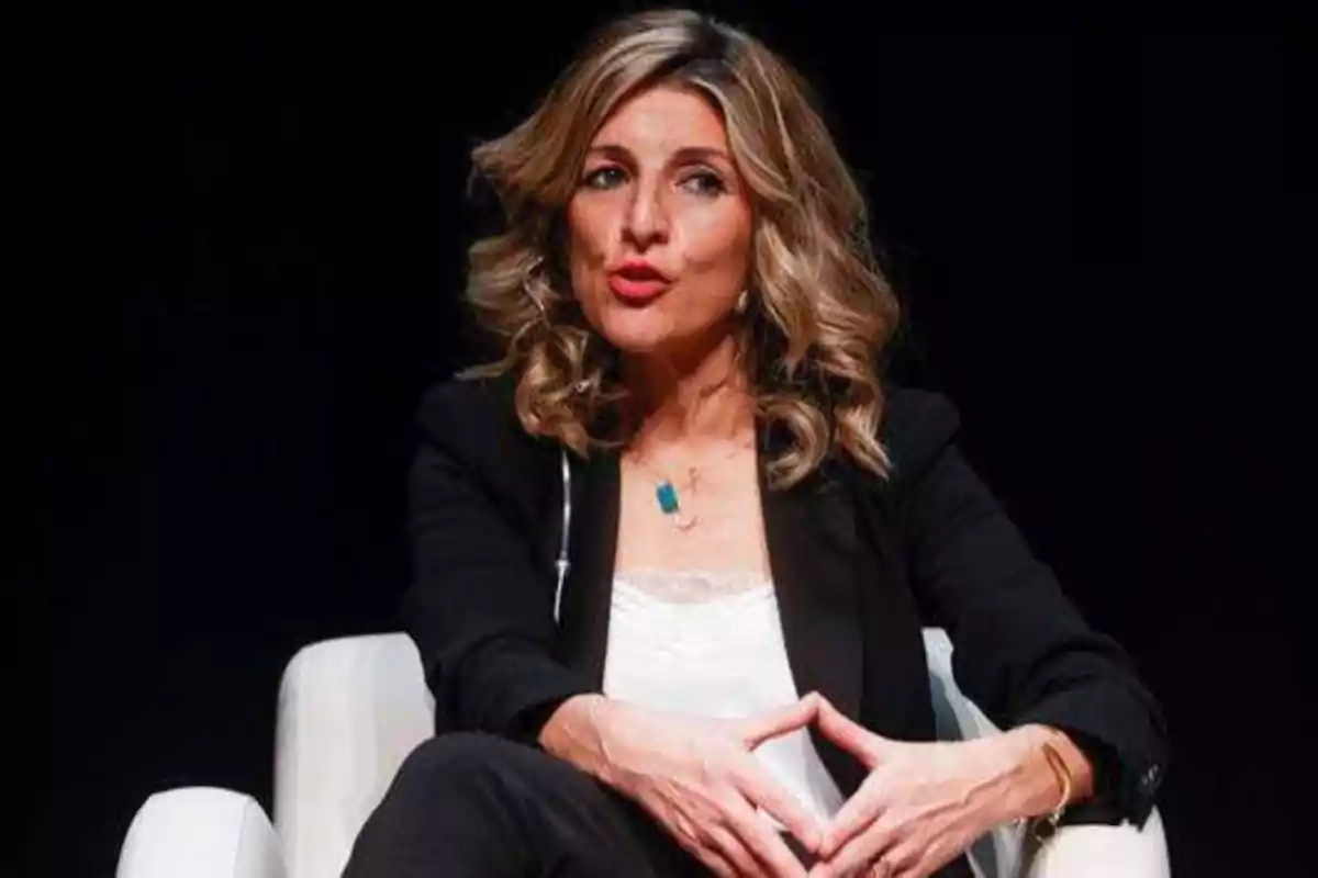 Una mujer con cabello rubio rizado y suelto, vestida con un blazer negro y una blusa blanca, está sentada en una silla blanca con las manos entrelazadas frente a ella, hablando en un entorno oscuro.