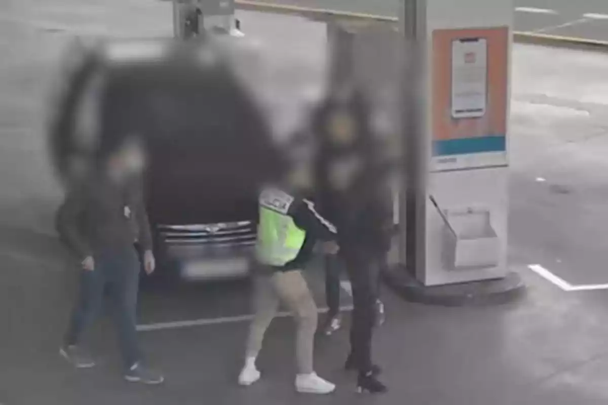 En la imagen se observa a varias personas en una estación de servicio, una de ellas parece estar siendo detenida por un individuo con un chaleco reflectante.