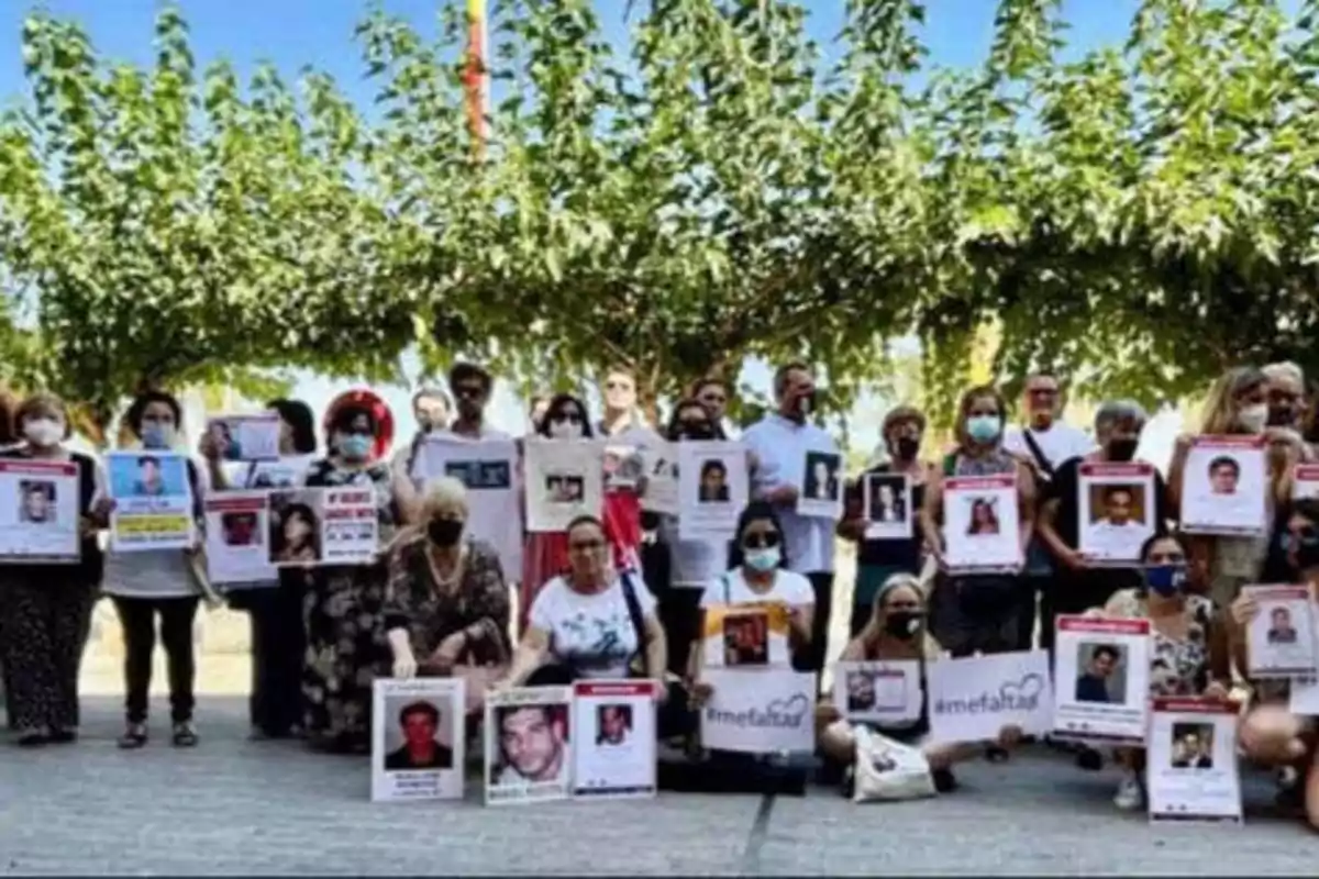 Un grupo de personas sosteniendo carteles con fotografías de individuos desaparecidos, reunidos al aire libre bajo árboles.