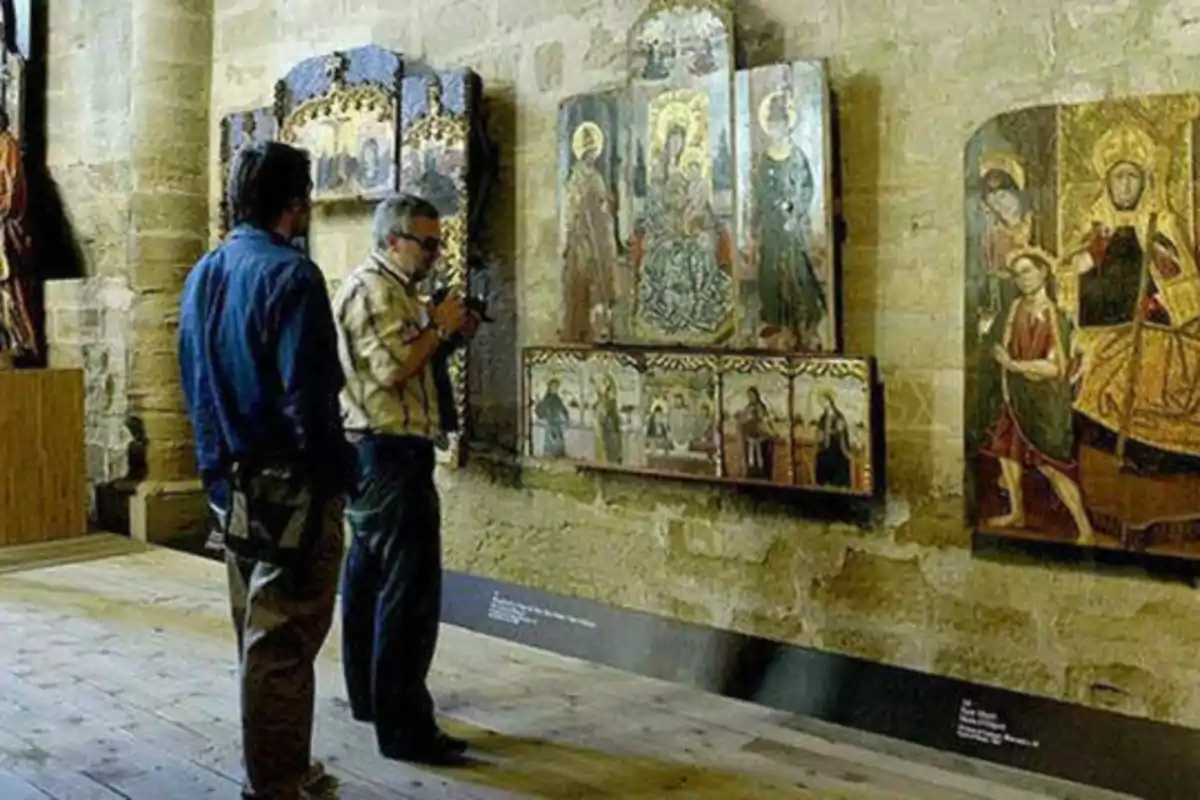 Dos personas observan pinturas religiosas en una galería de arte con paredes de piedra.