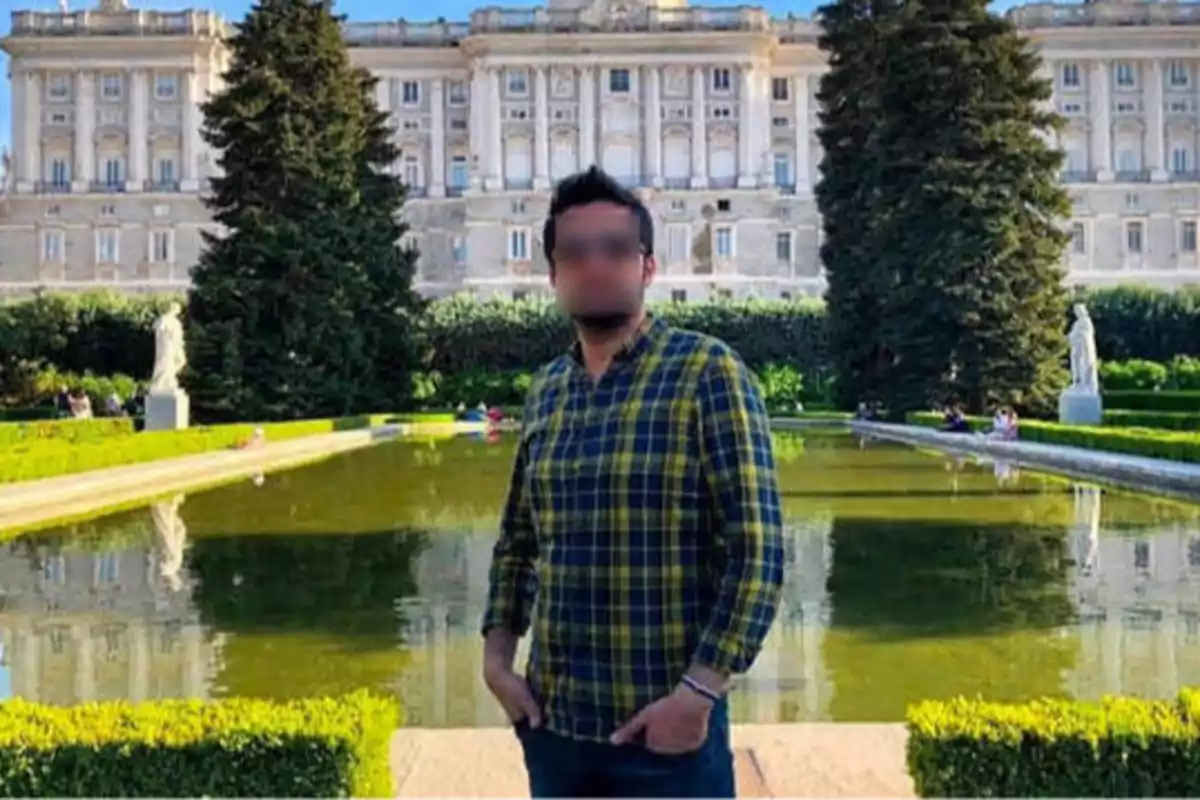 Persona con camisa de cuadros posando frente a un estanque en un jardín con un edificio histórico al fondo.