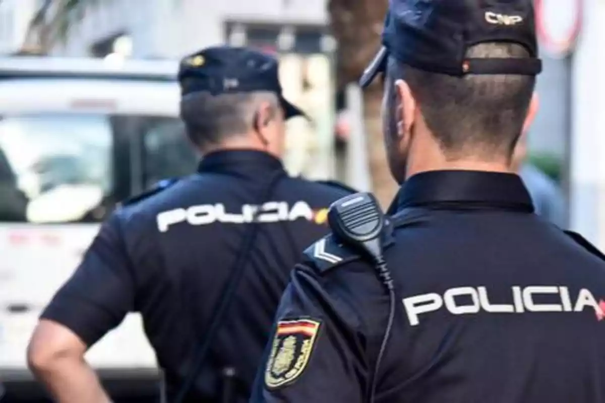 Dos policías de espaldas con uniformes y gorras, uno de ellos lleva una radio en el hombro.