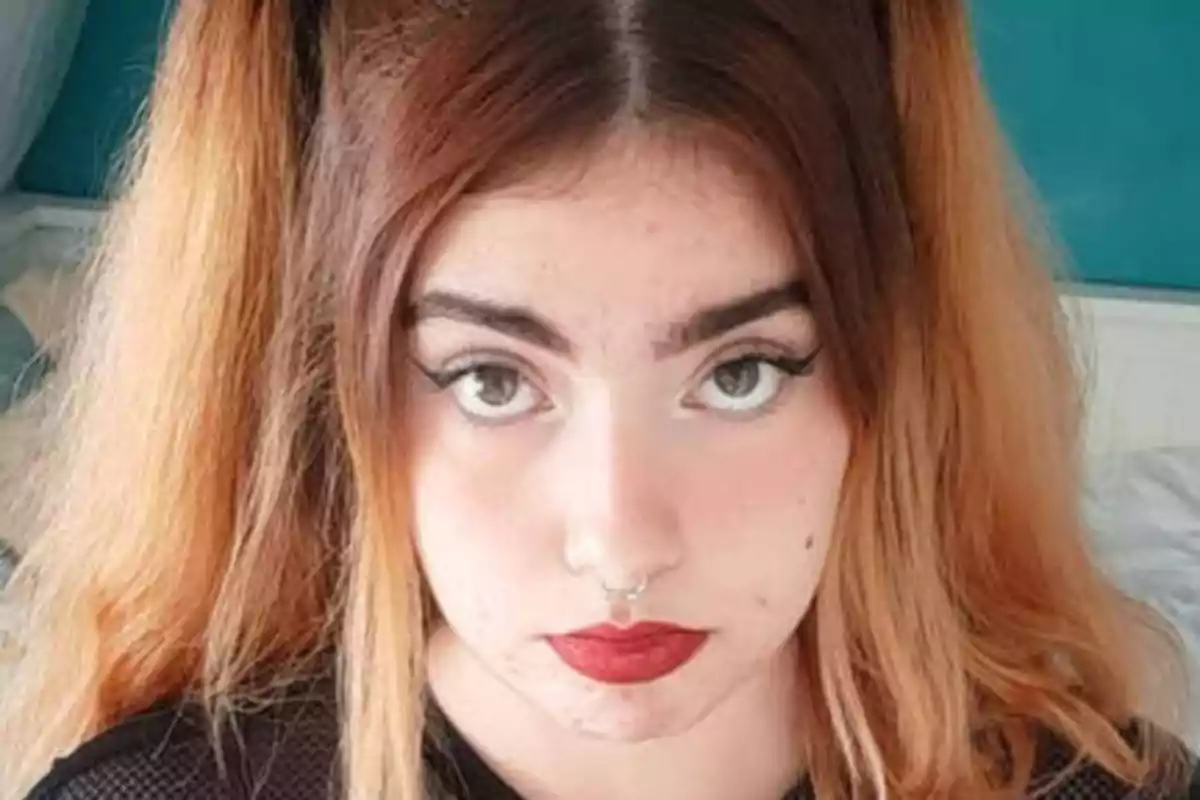 Una joven con cabello largo y teñido de rubio oscuro, con maquillaje en los ojos y labios rojos, mirando directamente a la cámara.