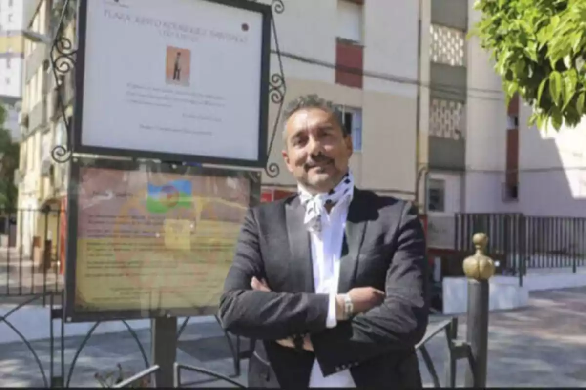 Un hombre con traje oscuro y camisa blanca, con una bufanda alrededor del cuello, está de pie con los brazos cruzados frente a un cartel informativo en una plaza urbana.