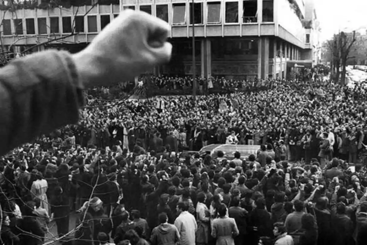 Una multitud de personas se reúne en una manifestación, con un puño levantado en primer plano y un edificio grande al fondo.