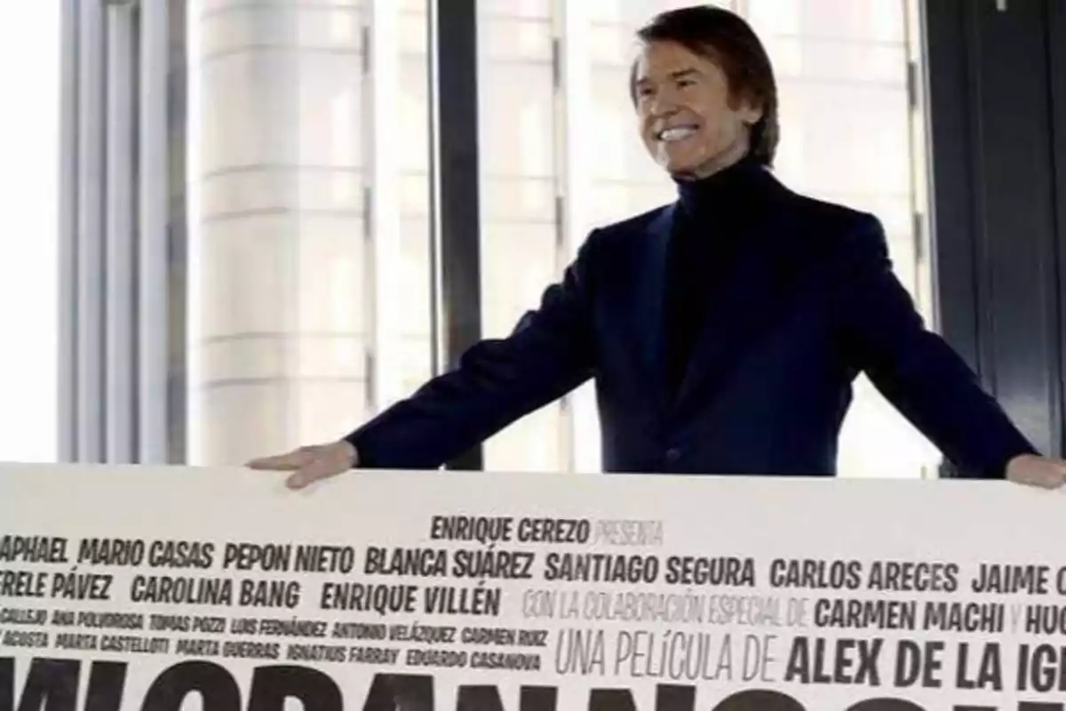 Un hombre sonriente sostiene un cartel promocional de una película con varios nombres de actores y actrices.