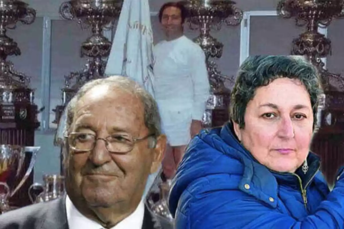 Tres personas posan frente a una colección de trofeos, una de ellas en el fondo con un uniforme blanco y las otras dos en primer plano, un hombre mayor con gafas y una mujer con chaqueta azul.