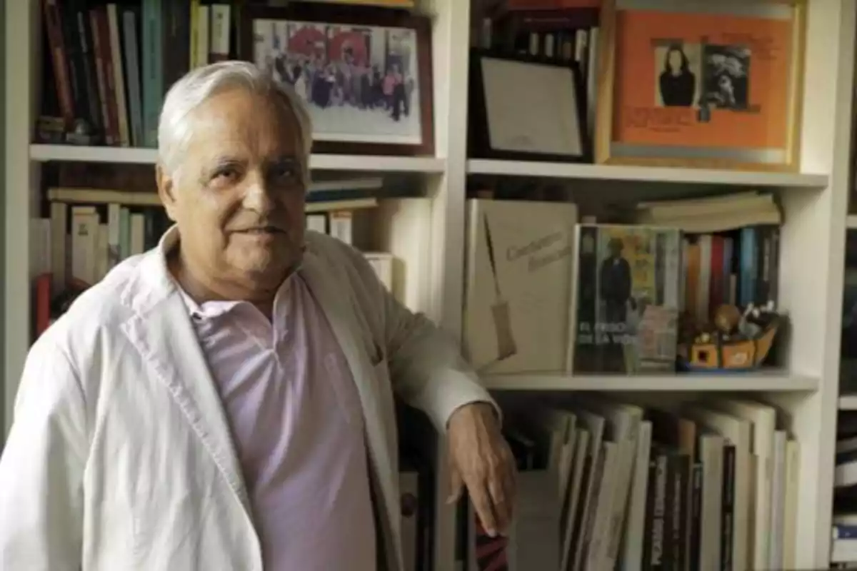 Un hombre de cabello canoso y chaqueta clara posa frente a una estantería llena de libros y fotografías.