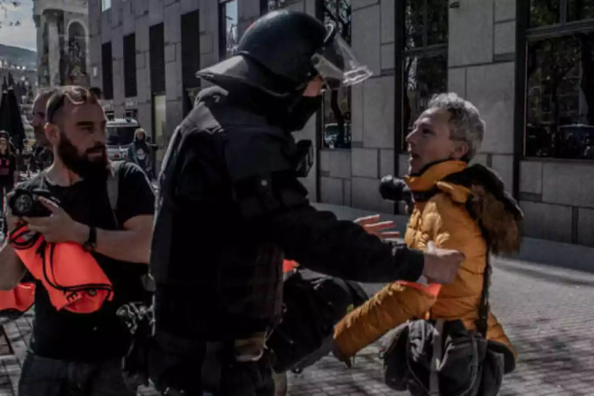 Un policía con equipo antidisturbios detiene a una persona con chaqueta amarilla mientras otra persona con una cámara observa la escena en una calle urbana.