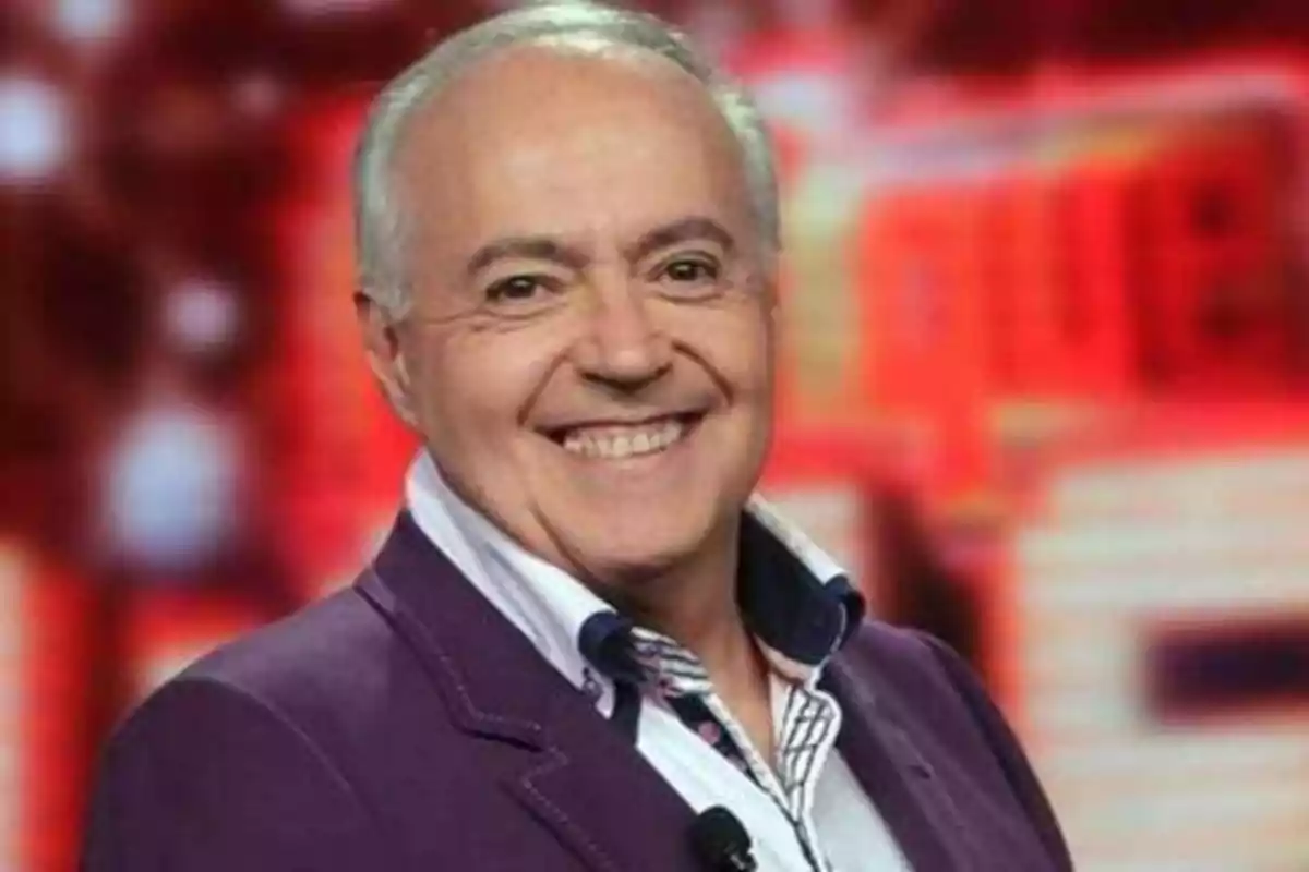 Un hombre mayor sonriente con cabello canoso y chaqueta morada frente a un fondo desenfocado de colores rojos y naranjas.