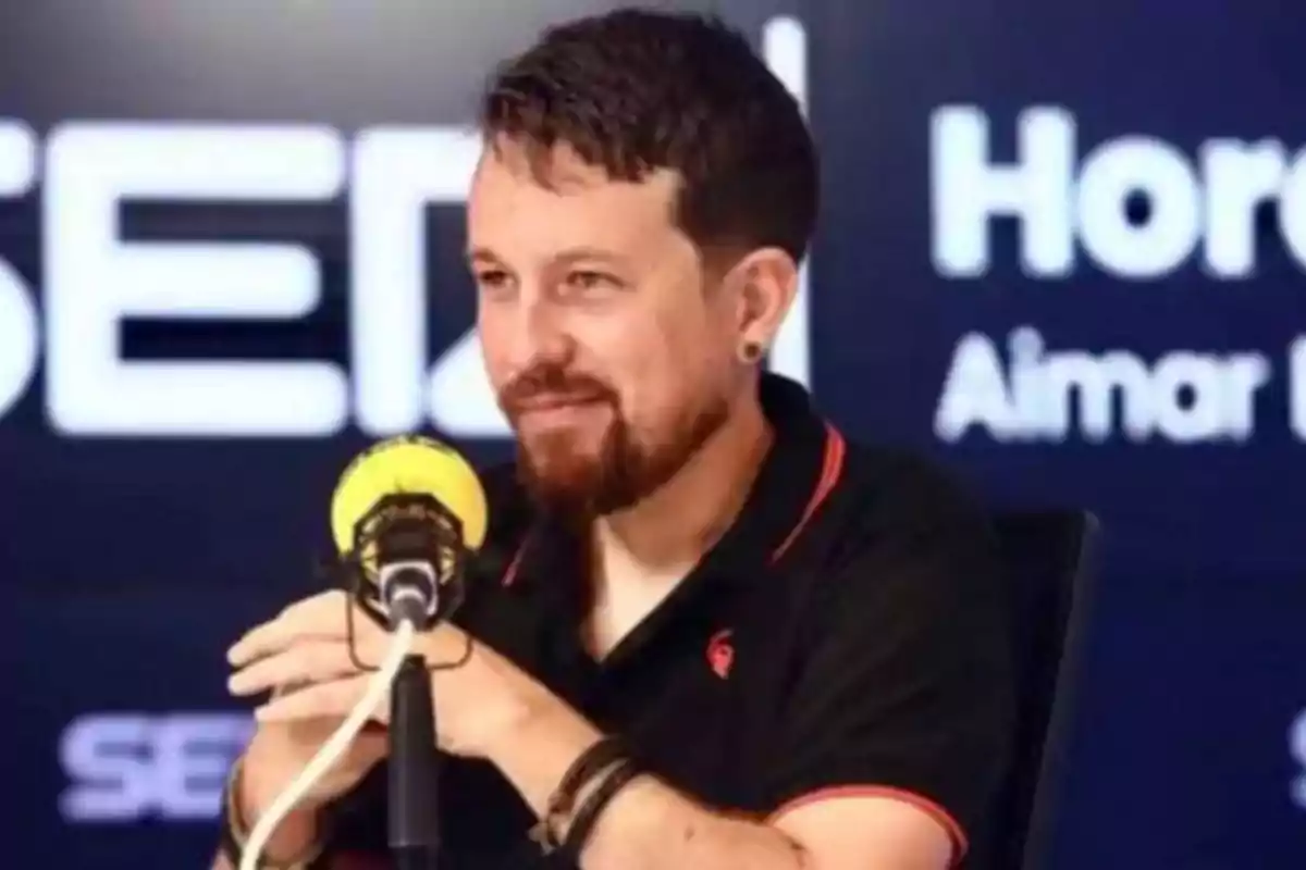 Un hombre con barba y cabello corto está hablando en un micrófono amarillo en un estudio de radio con el logo de la cadena SER en el fondo.