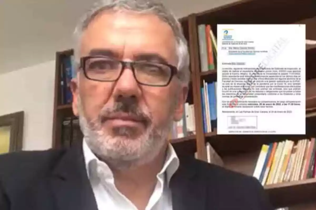 Un hombre con gafas y barba gris frente a una estantería con libros, con un documento visible en la esquina superior derecha.