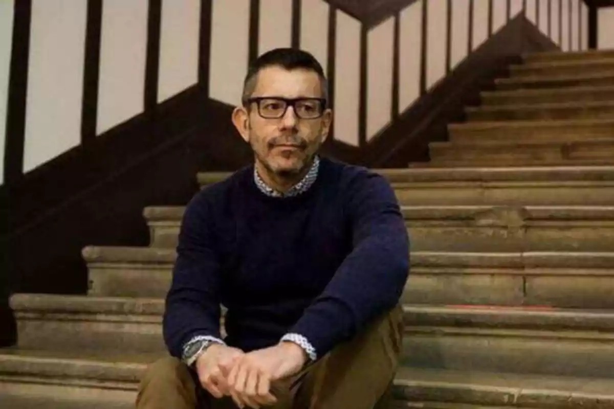 Hombre con gafas y barba sentado en una escalera de madera con barandales blancos y negros.