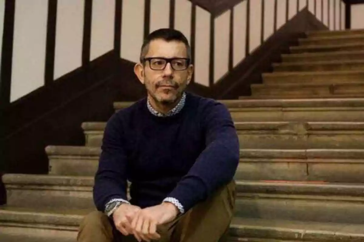 Hombre con gafas y barba sentado en una escalera de madera con barandales blancos y negros.