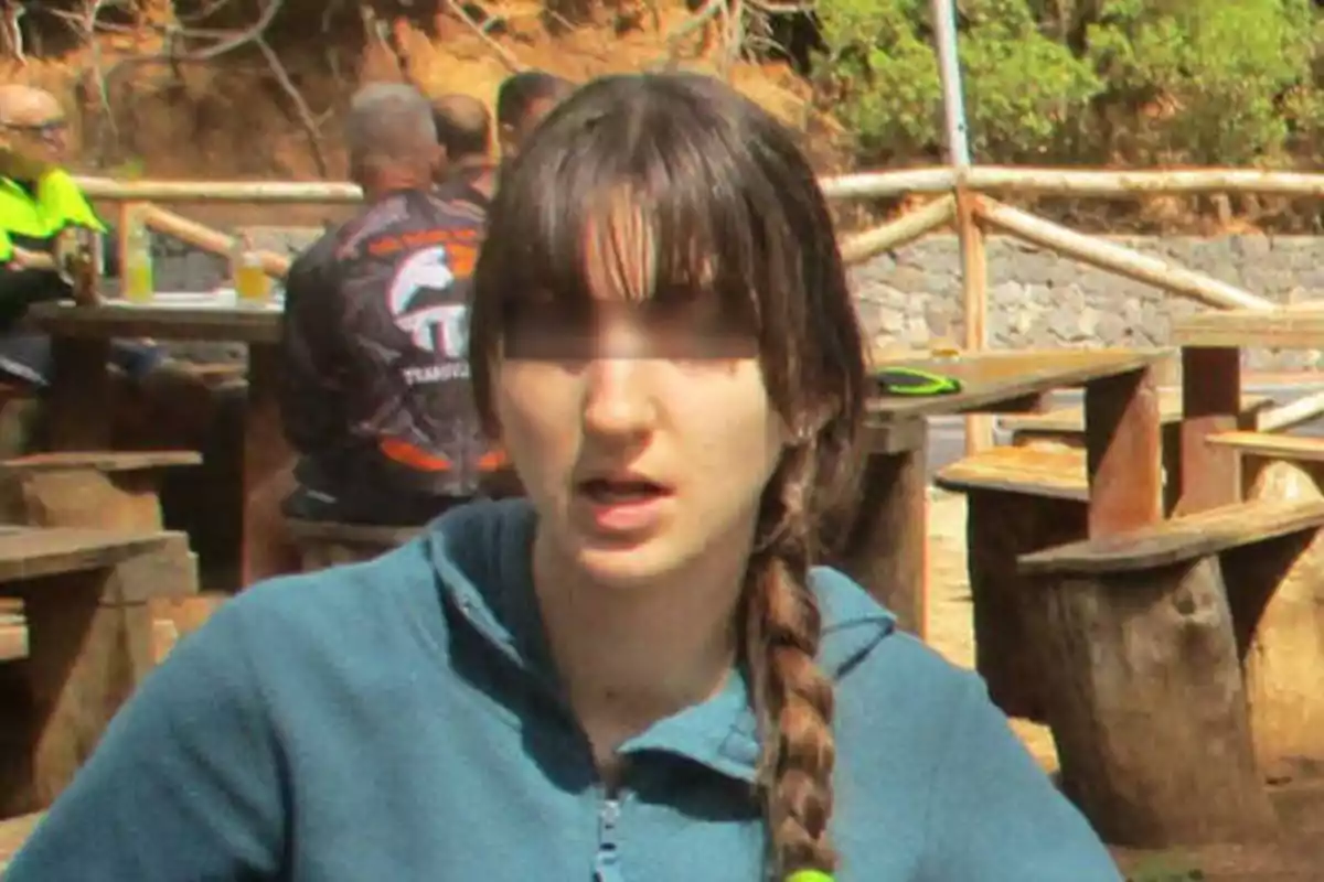 Una persona con una trenza y una sudadera azul está sentada en un área al aire libre con mesas de madera y otras personas al fondo.