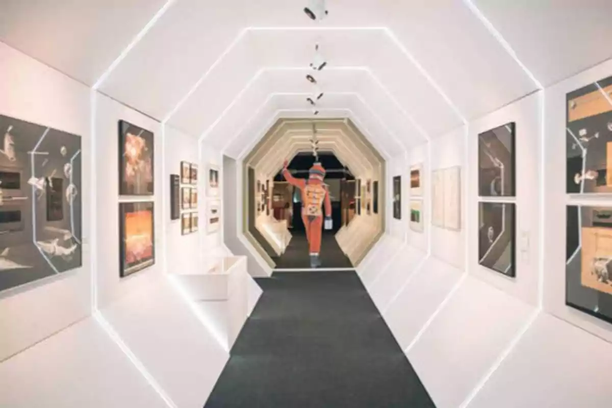 Un pasillo futurista con paredes blancas y luces LED, decorado con cuadros y fotografías, y una figura humana al fondo.