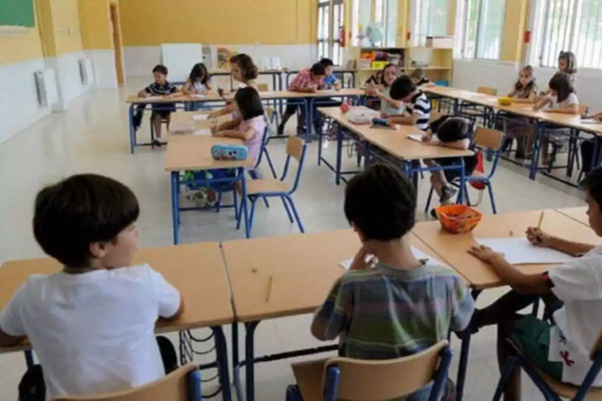Niños sentados en escritorios dentro de un aula escolar, algunos escribiendo y otros conversando.