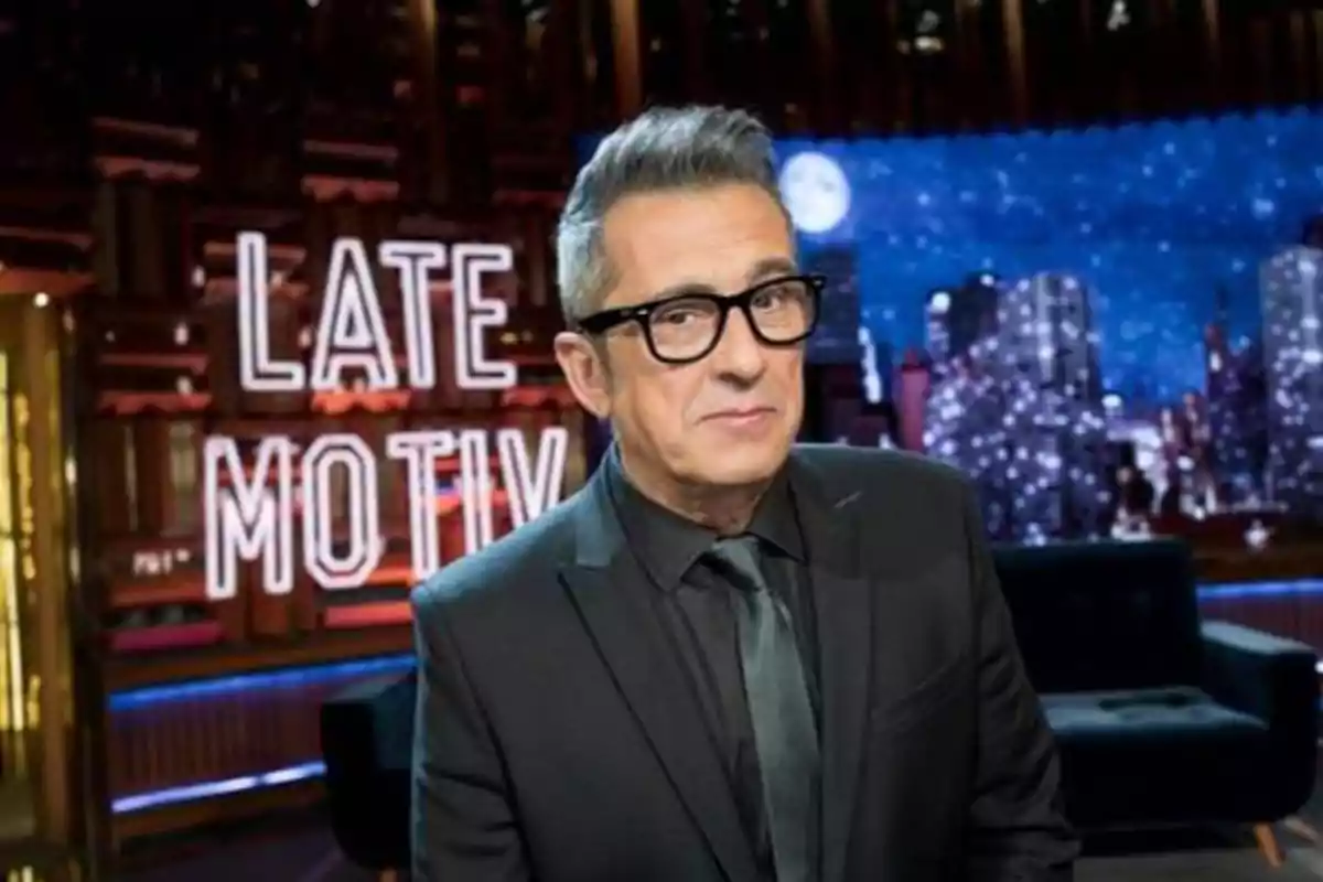 Hombre con gafas y traje oscuro en un set de televisión con el letrero "Late Motiv" iluminado en el fondo.