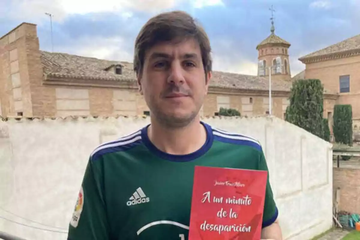 Hombre con camiseta verde sosteniendo un libro rojo frente a un edificio de ladrillo.