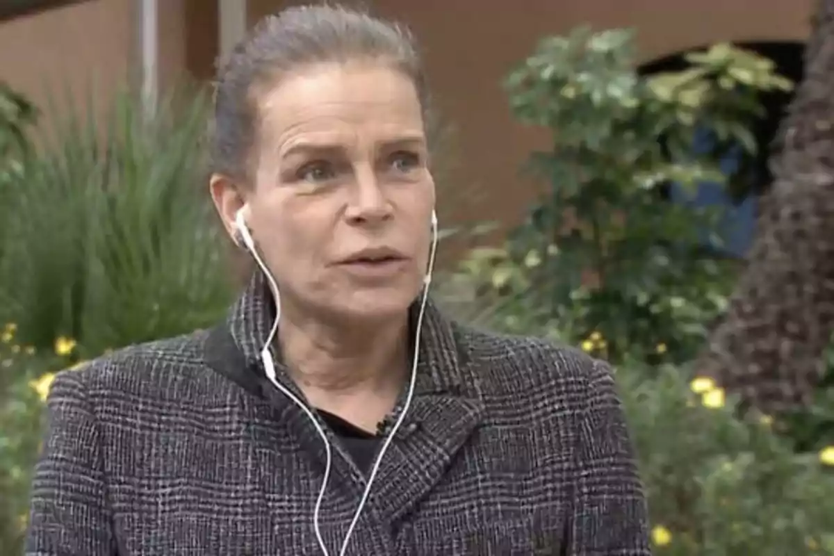 Una mujer con auriculares blancos y chaqueta gris, hablando al aire libre con plantas de fondo.