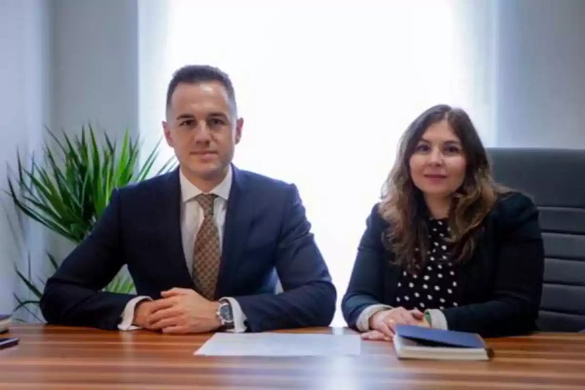 Dos personas sentadas en una mesa de oficina con una planta en el fondo.