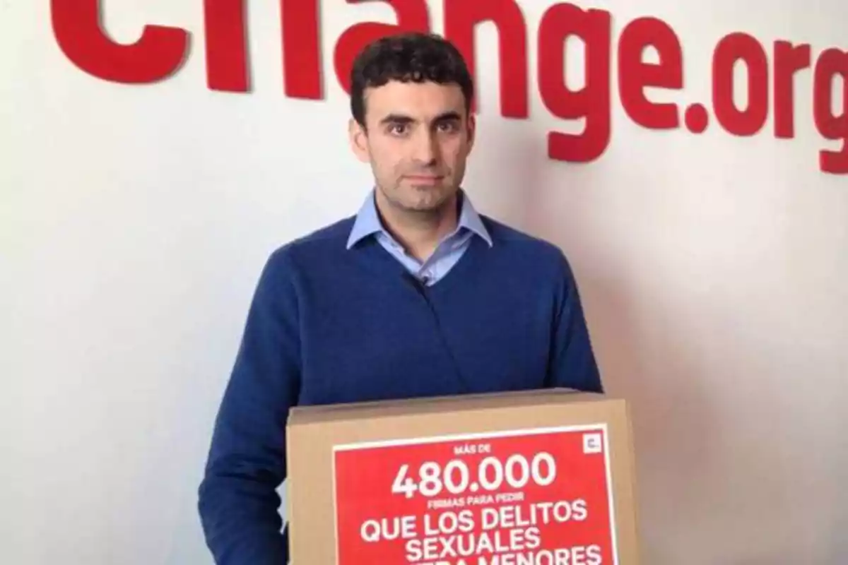 Un hombre sosteniendo una caja con un cartel que dice "Más de 480,000 firmas para pedir que los delitos sexuales a menores no prescriban" frente a un fondo con el logo de Change.org.