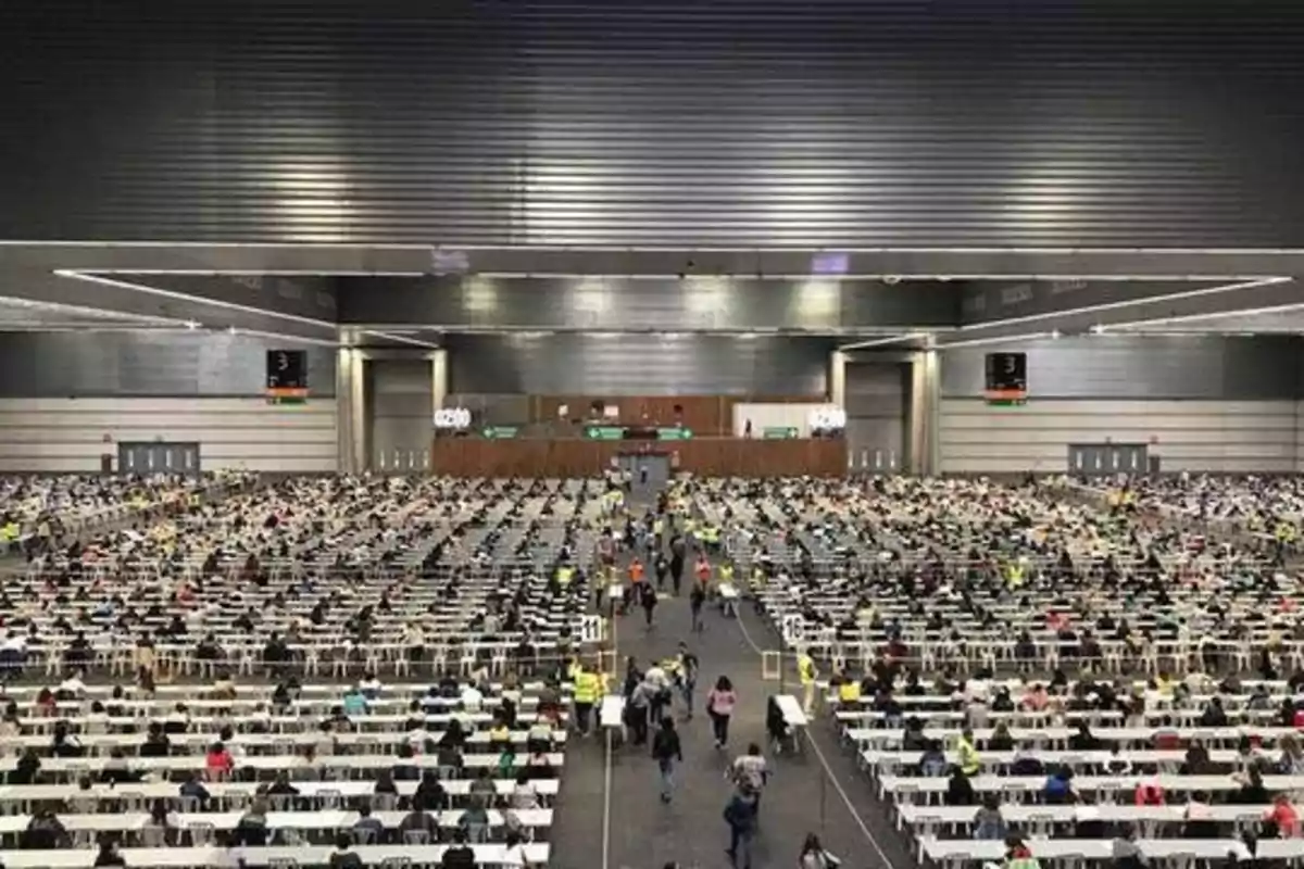 Una gran sala llena de personas sentadas en mesas largas, posiblemente participando en un examen o evento masivo.