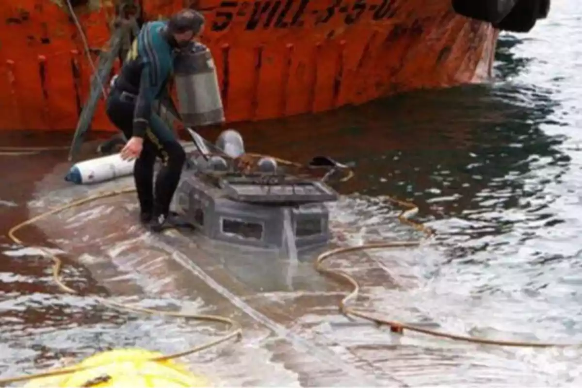 Un buzo inspecciona un pequeño submarino parcialmente sumergido junto a un barco naranja.