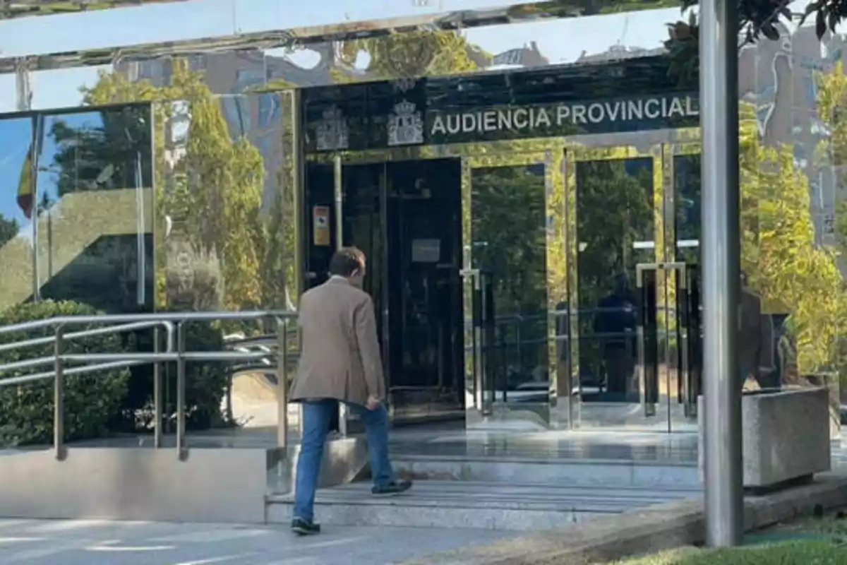 Un hombre con chaqueta marrón y jeans sube las escaleras hacia la entrada de la Audiencia Provincial, un edificio con fachada de vidrio reflectante y barandillas metálicas.