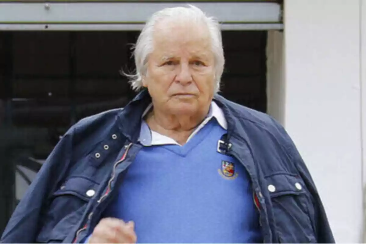 Un hombre mayor con cabello canoso y expresión seria, viste una chaqueta azul oscuro sobre un suéter azul claro con un escudo bordado, y una camisa blanca debajo.