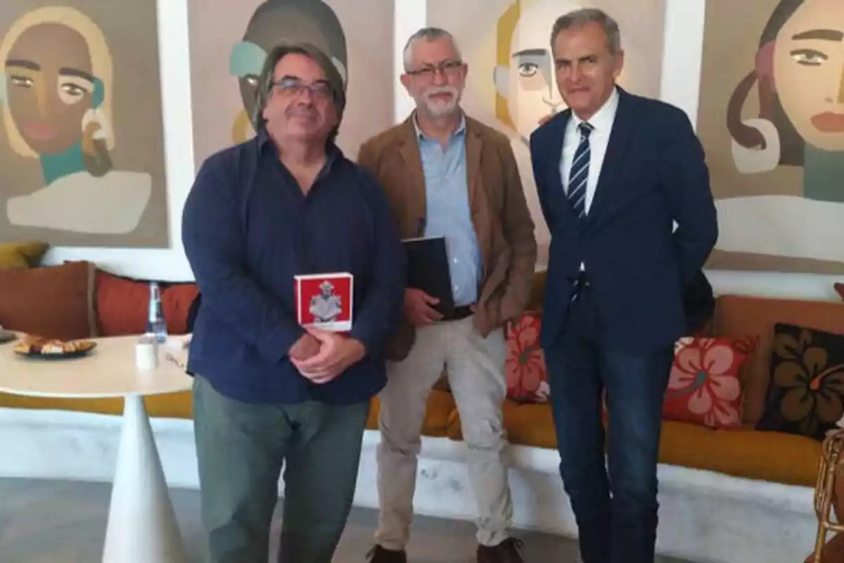 Tres hombres posan frente a una pared con retratos artísticos, uno de ellos sostiene un libro rojo.