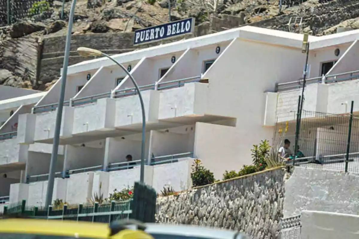 La imagen muestra un edificio de apartamentos blancos de dos pisos con balcones, ubicado en una zona rocosa y con un letrero que dice "Puerto Bello" en la parte superior.