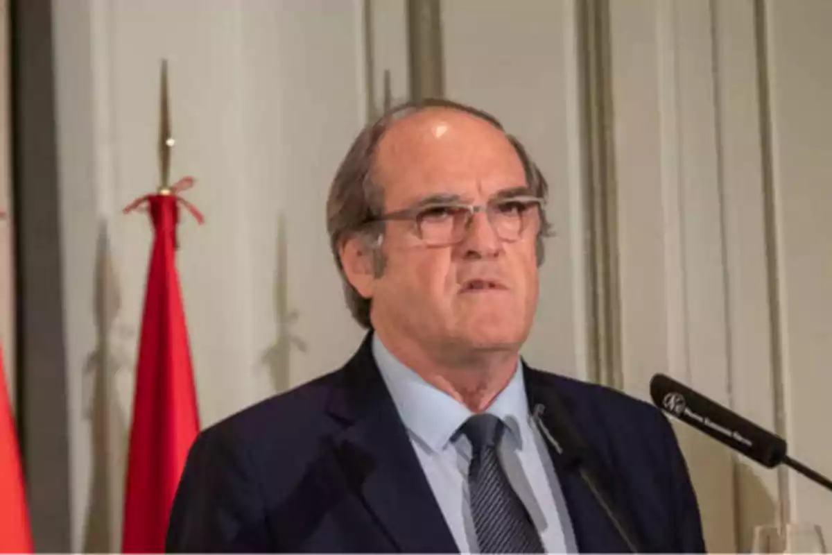 Hombre de mediana edad con gafas y traje oscuro hablando en un micrófono con una bandera roja de fondo.