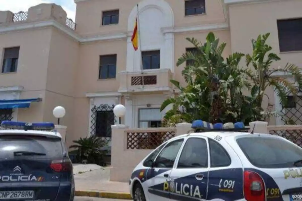 Edificio de una comisaría de policía con dos coches patrulla estacionados al frente y una bandera ondeando en la fachada.