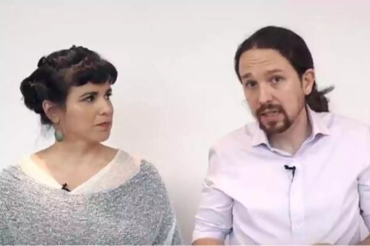 Dos personas sentadas, una mujer con cabello oscuro y una blusa gris, y un hombre con barba y cabello largo, ambos con micrófonos.