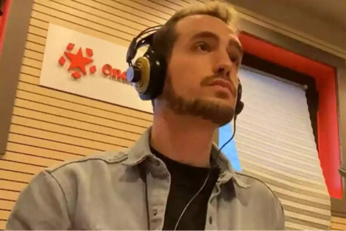 Un hombre con auriculares y chaqueta de mezclilla en un estudio de radio con un letrero de fondo.