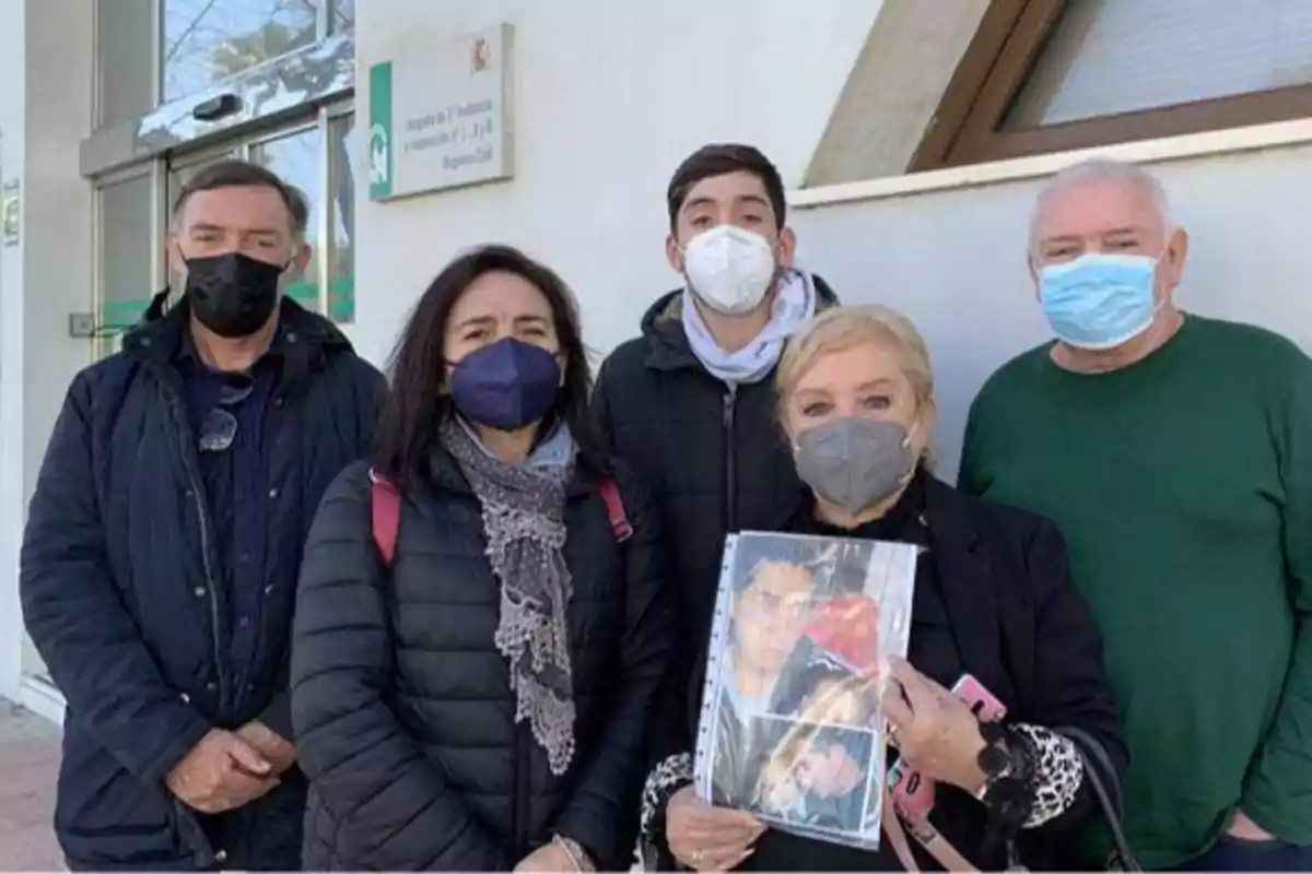 Un grupo de personas con mascarillas posan frente a un edificio, una de ellas sostiene una carpeta con fotografías.